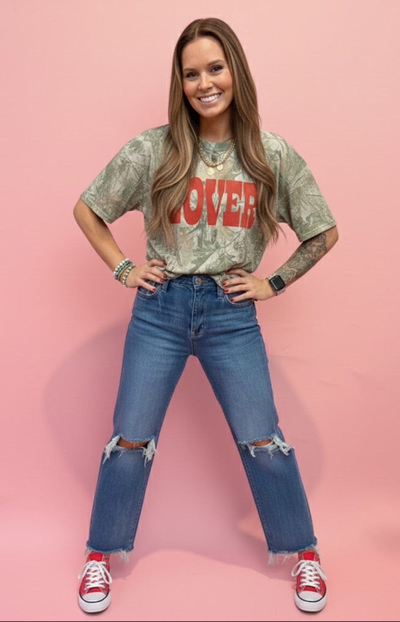 woman standing in front of pink wall wearing camo tshirt and jeans.