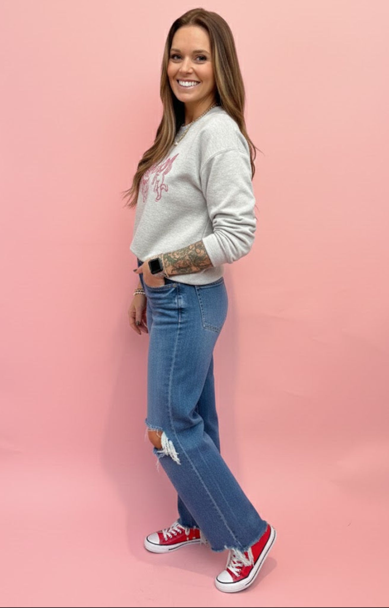 woman wearing graphic sweatshirt with cupids and jeans with red sneakers on a pink background