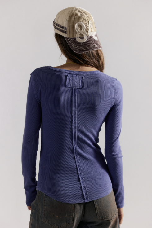 A woman shown from the back wearing a textured, long-sleeve ribbed knit top in a muted purple-blue shade, paired with dark gray pants and a beige baseball cap.