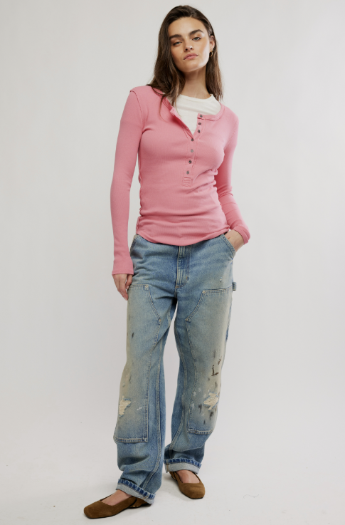 A woman modeling a pink long-sleeve henley top with a white undershirt, distressed light blue jeans, and brown flats, standing against a plain white studio background.